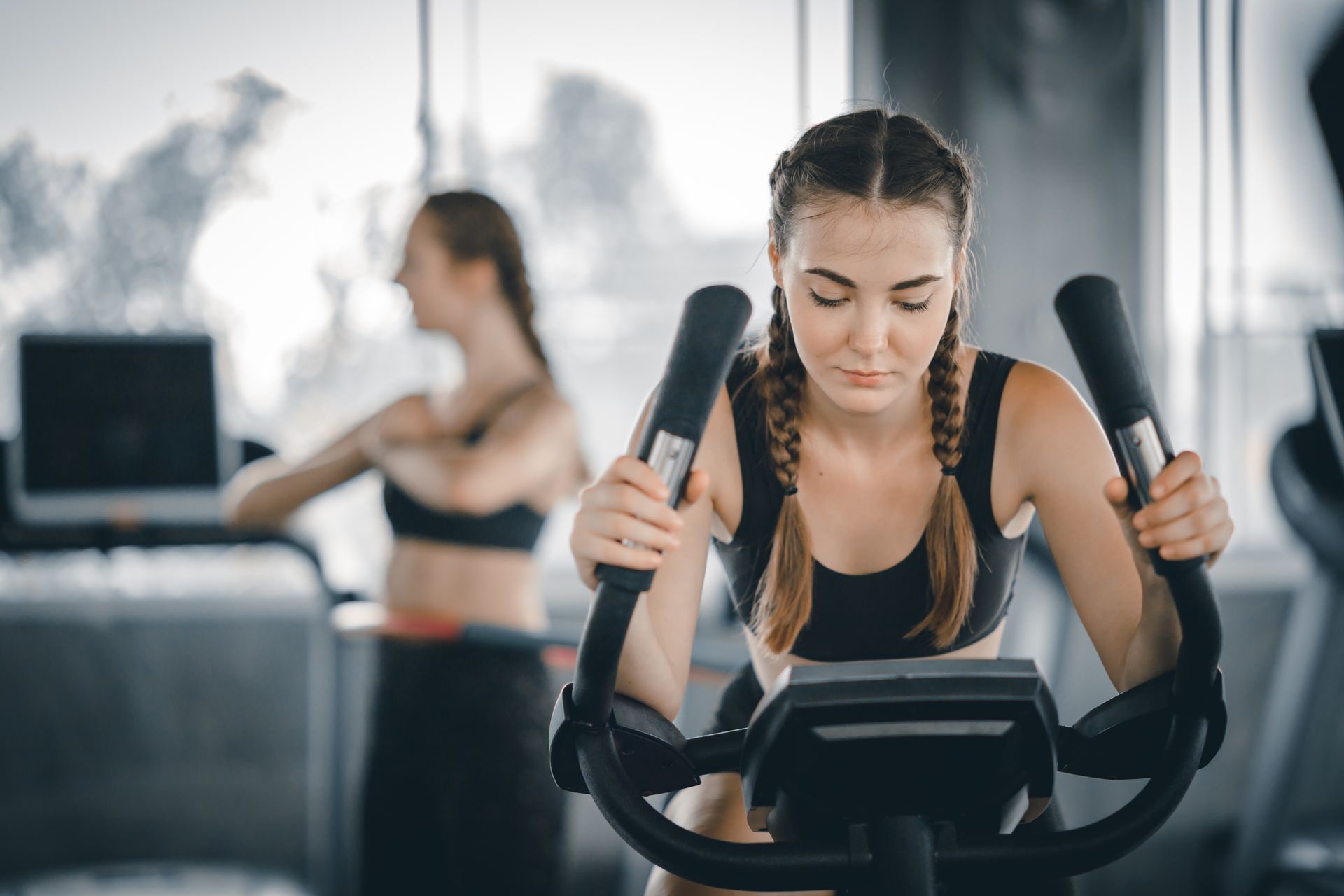 Mulher se exercitando em uma máquina elíptica na academia; outra mulher se alongando ao fundo.