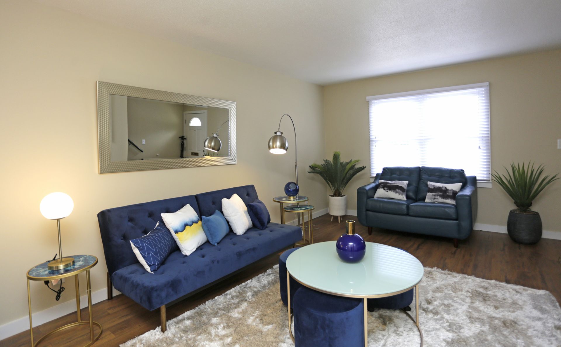 Blue velvet sofa and loveseat in a living room with a round coffee table, a mirror, and potted plants.