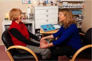 Two women sit facing each other in a clinic, holding hands during a conversation.