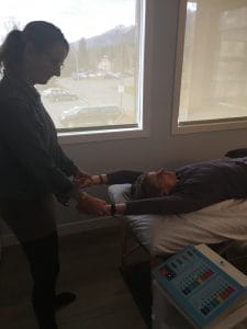Person standing beside a massage table, holding hands with a person lying down by a window.