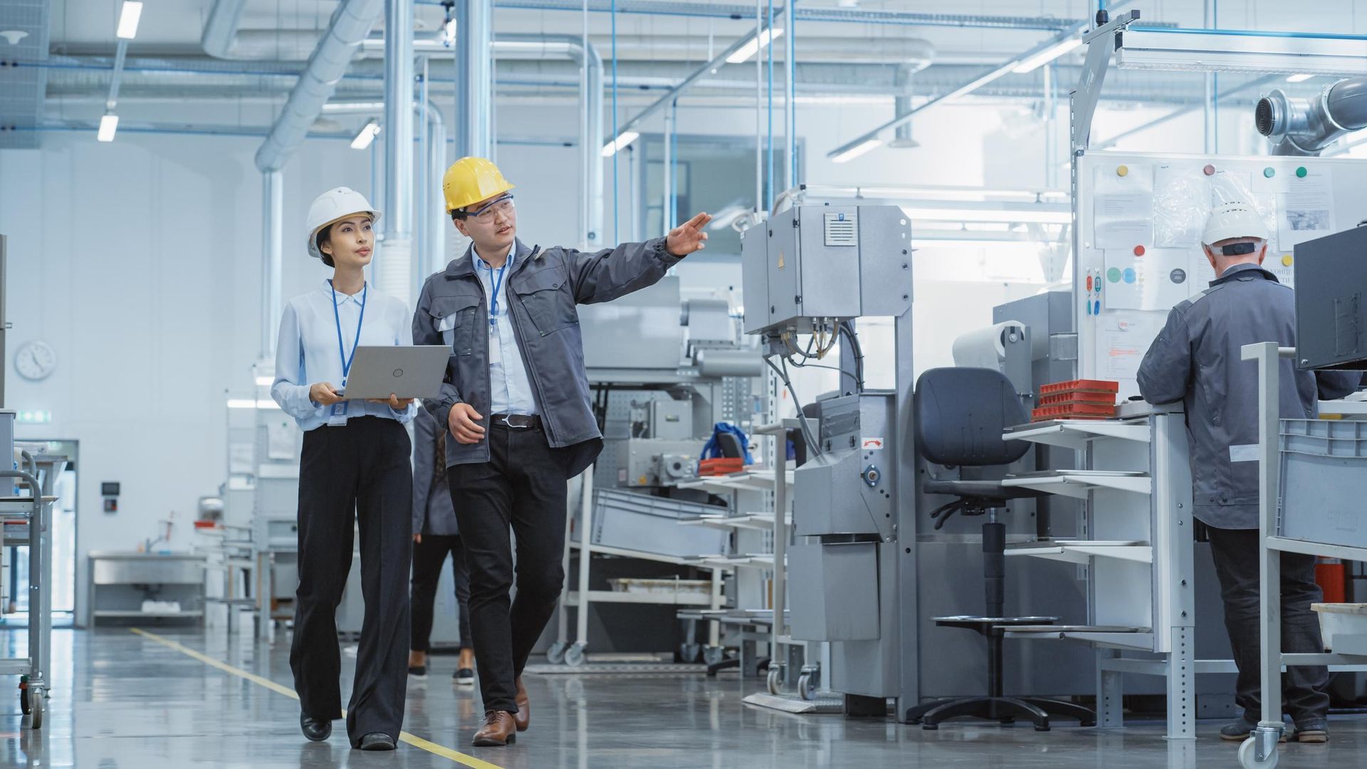 Two engineers in hard hats inspect factory machinery in a bright industrial workshop.