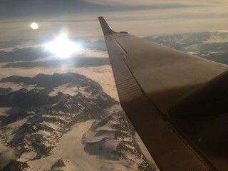 Airplane wing over snowy mountains and clouds at sunset with bright sun reflecting on water