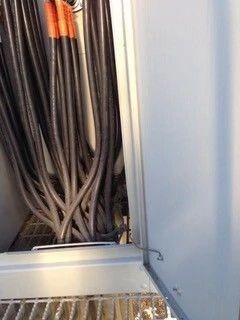 Bundle of thick black cables in a utility cabinet, with gray metal panels and orange cable ends at top.