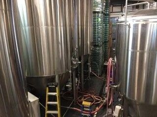 Stainless steel brewery tanks with a yellow ladder and hoses in a utility room