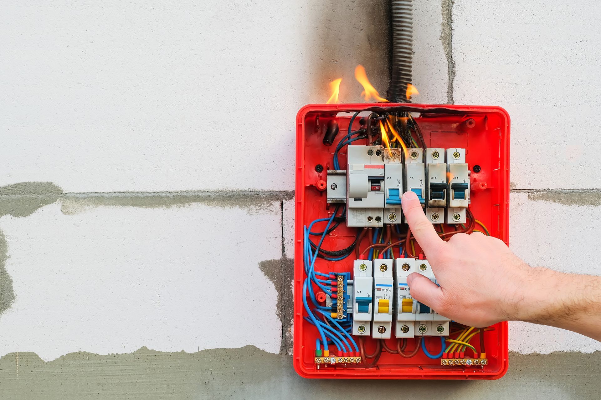 Hand pointing at a smoking red electrical breaker panel with exposed wires on a wall