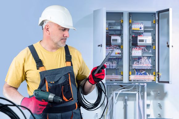 Technician in hard hat inspecting an open electrical control panel with testing tools