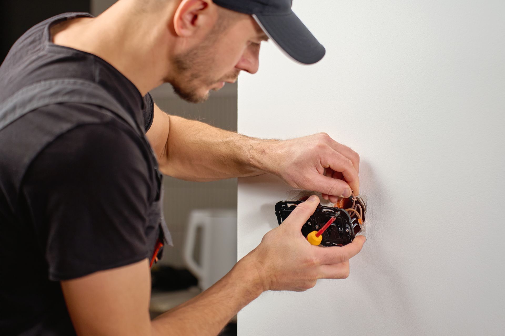 Person in a cap working on a wall-mounted device with exposed wires in a home interior