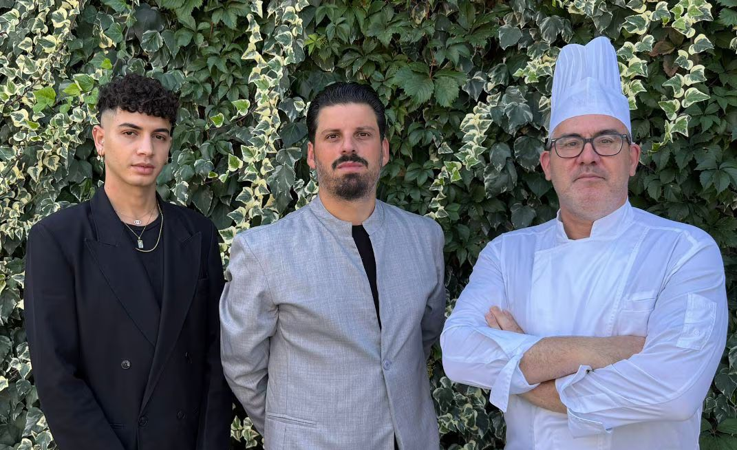 Tre persone in piedi all'aperto davanti a un muro ricoperto di foglie, una delle quali con un cappello da chef