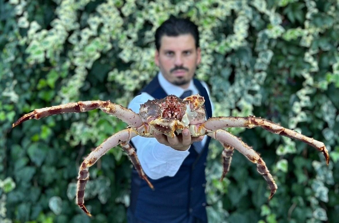 Un uomo tiene in mano un ragno gigante simile a un granchio, rivolto verso la telecamera, con del fogliame verde sullo sfondo.