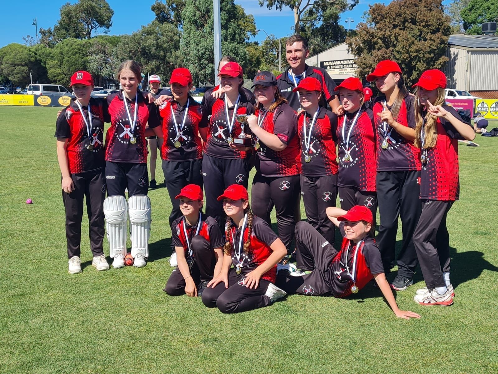Youth girls cricket team in red-and-black uniforms posing on a sunny field with trophies and medals
