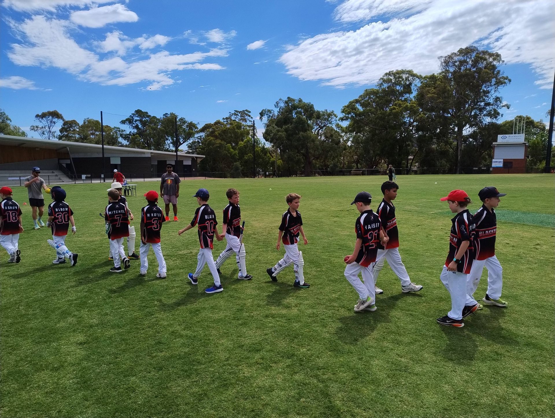 Children in black jerseys and white pants walk across a grassy field under a blue sky.