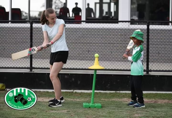 Cricket batter swings at a ball on a tee while a coach demonstrates next to her.