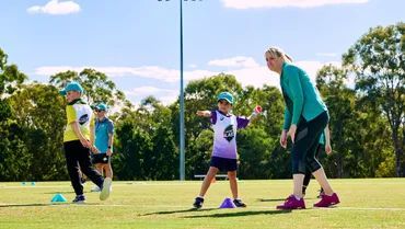 Coach instructs three children in cricket uniforms to throw a ball on a sunny grass field with cones.