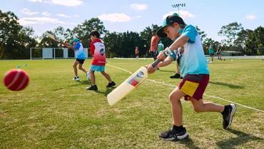 Children playing cricket on a sunny field, with one boy swinging a bat at a red ball.