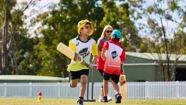 Three children play cricket on a grassy field, one in yellow batting while others watch nearby.