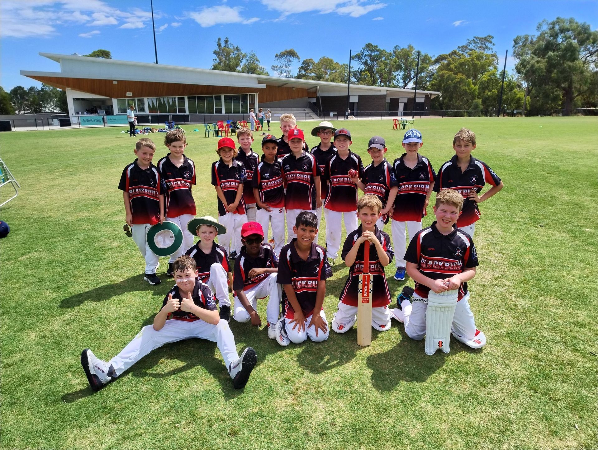 Youth cricket team posing on a grassy field in black uniforms, holding bats and medals, with clubhouse behind them
