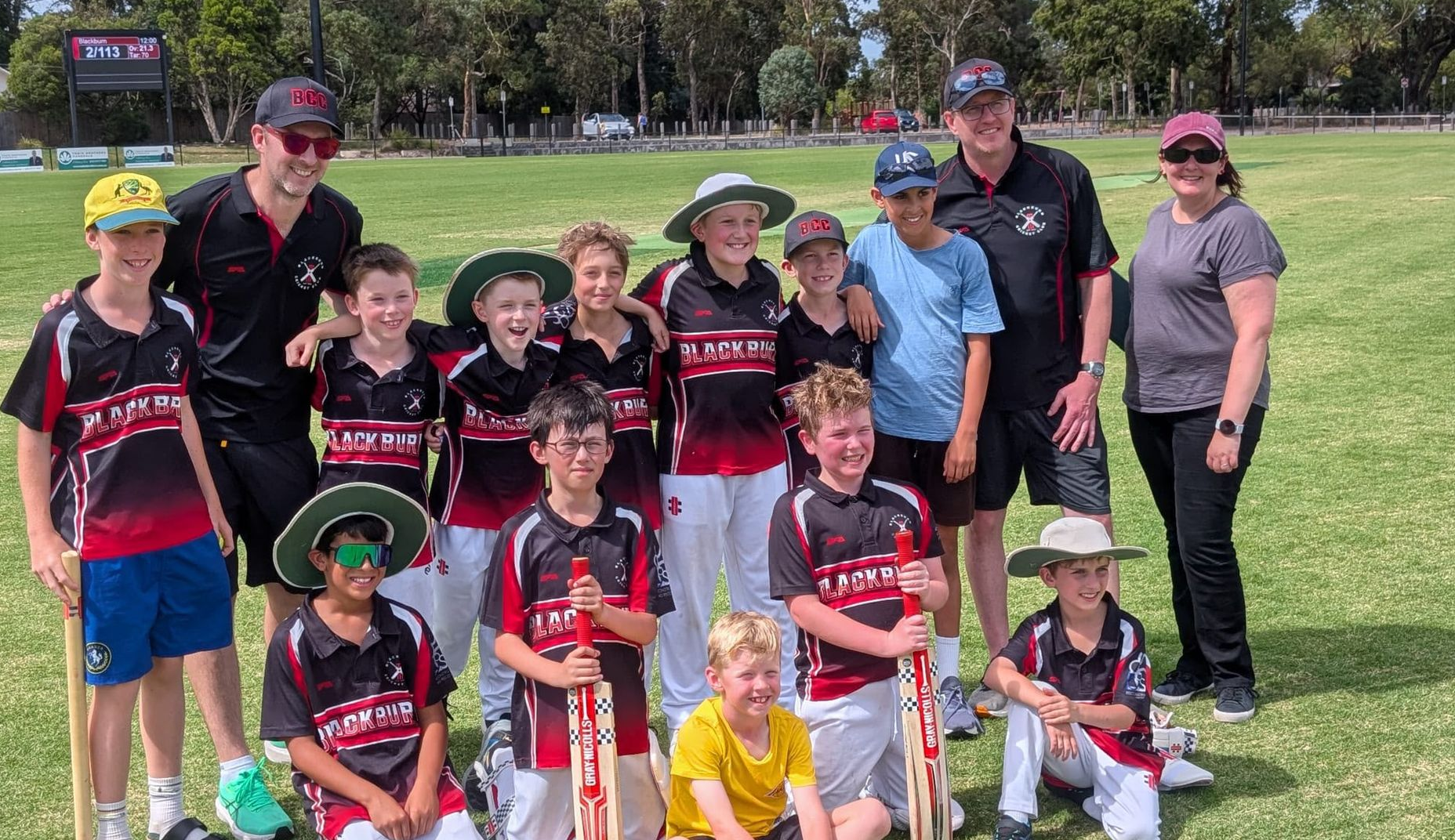 Youth cricket team posing on a grassy field, wearing red and black uniforms with coaches behind them.