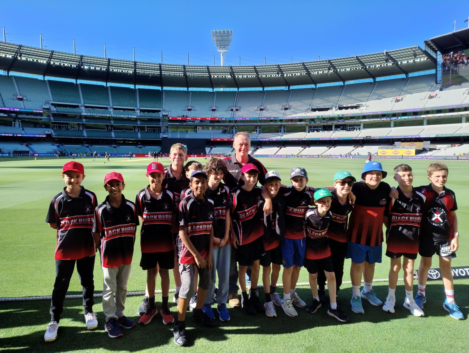 Children in black and red sports uniforms standing on a stadium field with a coach behind them