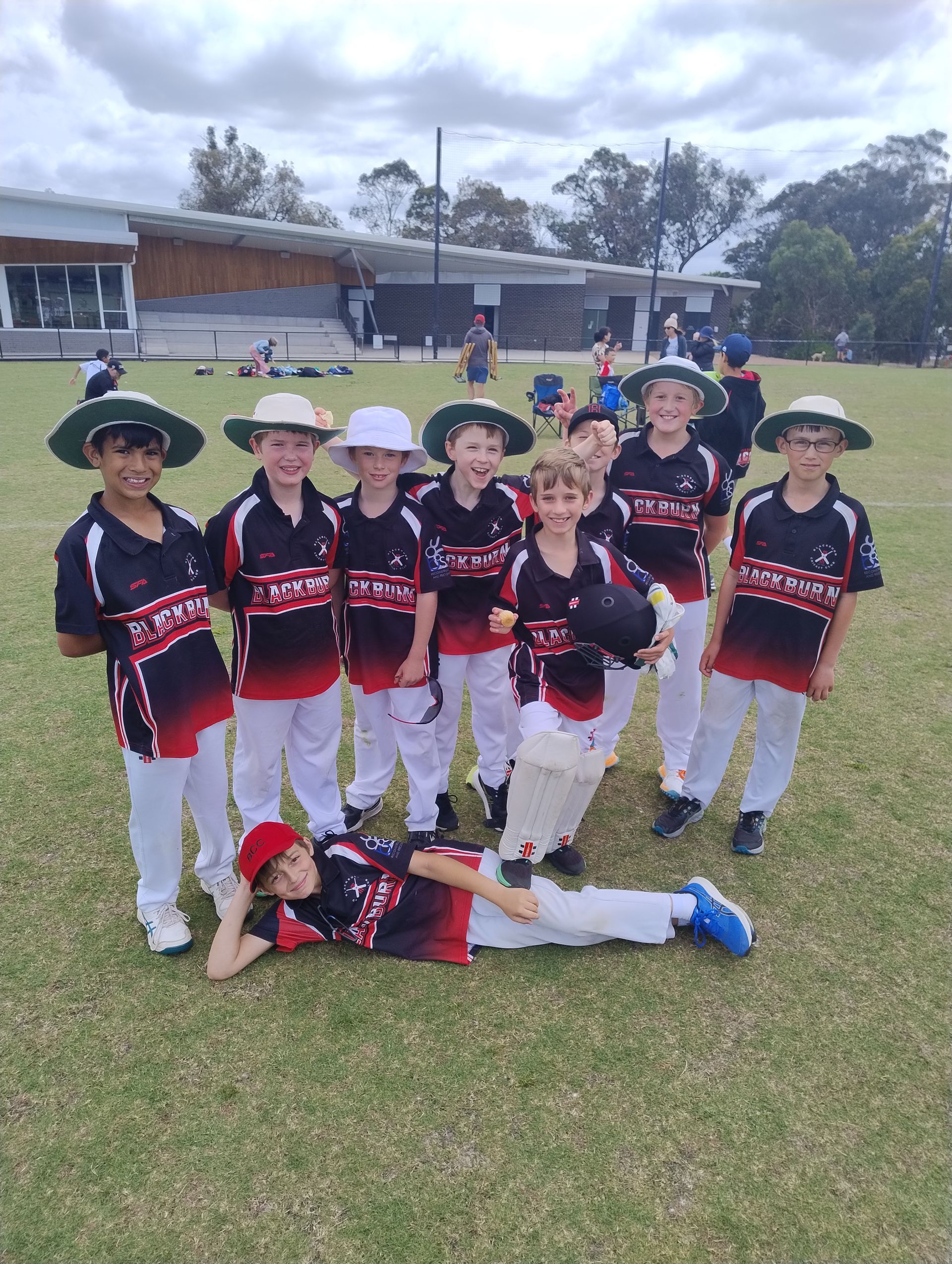 Youth cricket team in black, red, and white uniforms posing on a grassy field with one child lying in front.