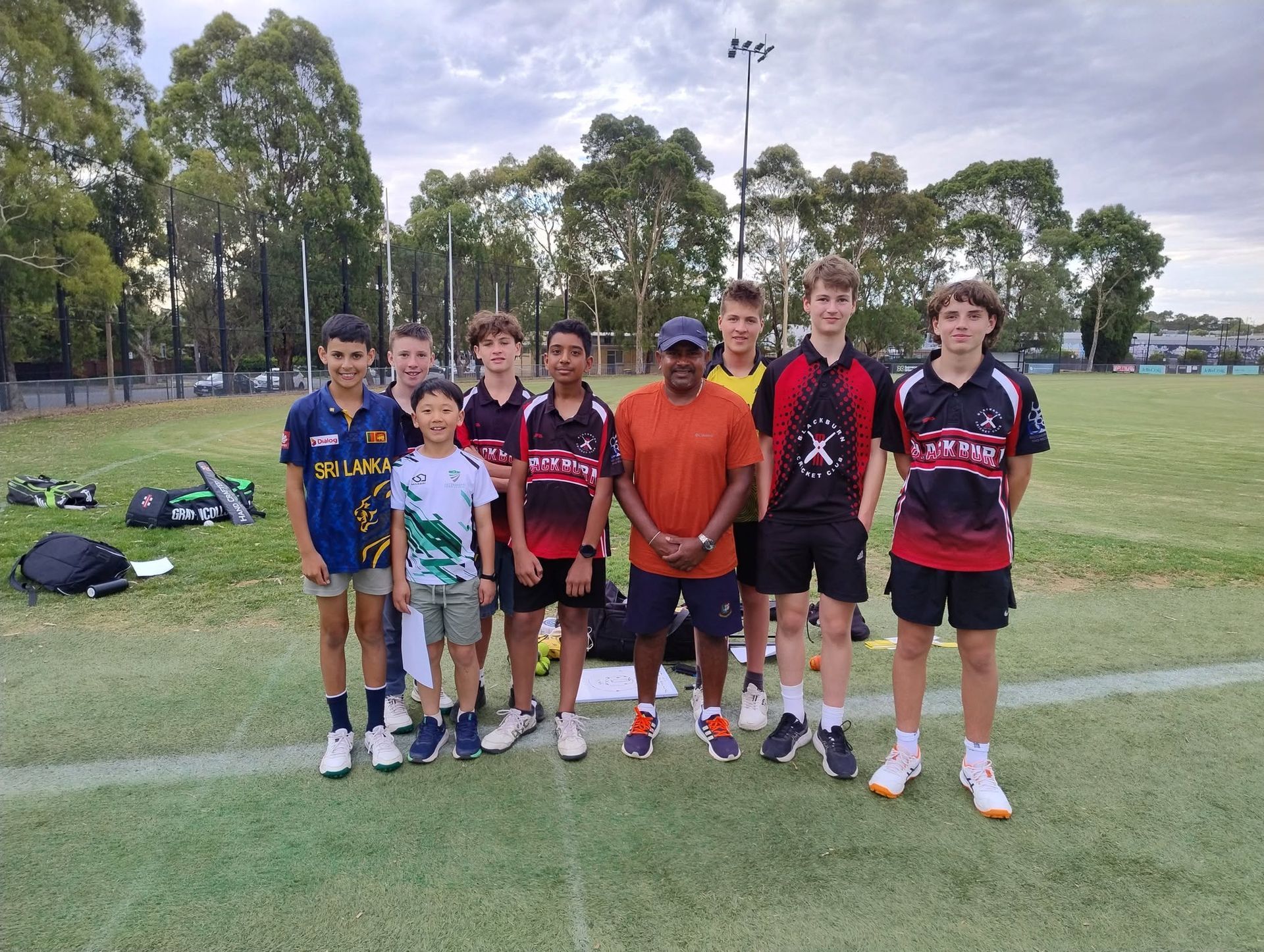 Seven young cricketers posing on a grassy sports field with Sri Lankan cricketer Rangana Herath