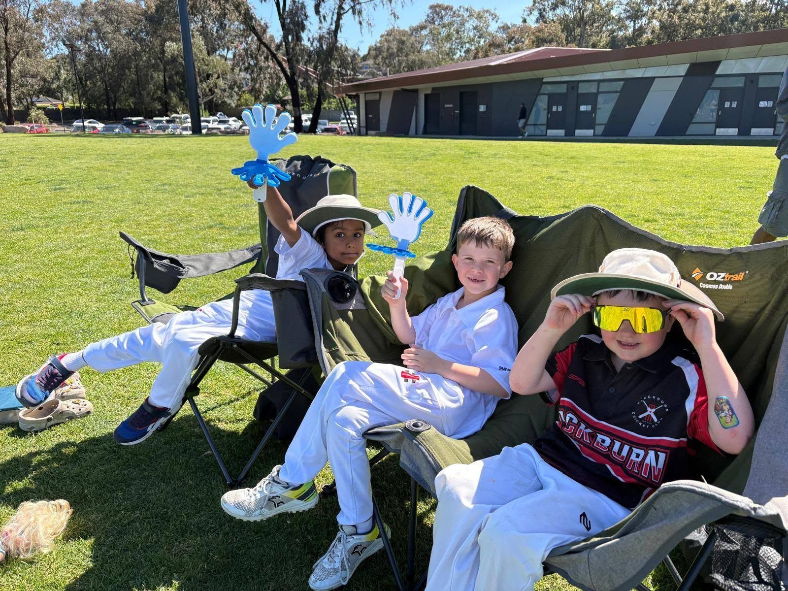 Three juniors lounge on chairs on a sunny field, smiling and waving blue paper hands near a school building.