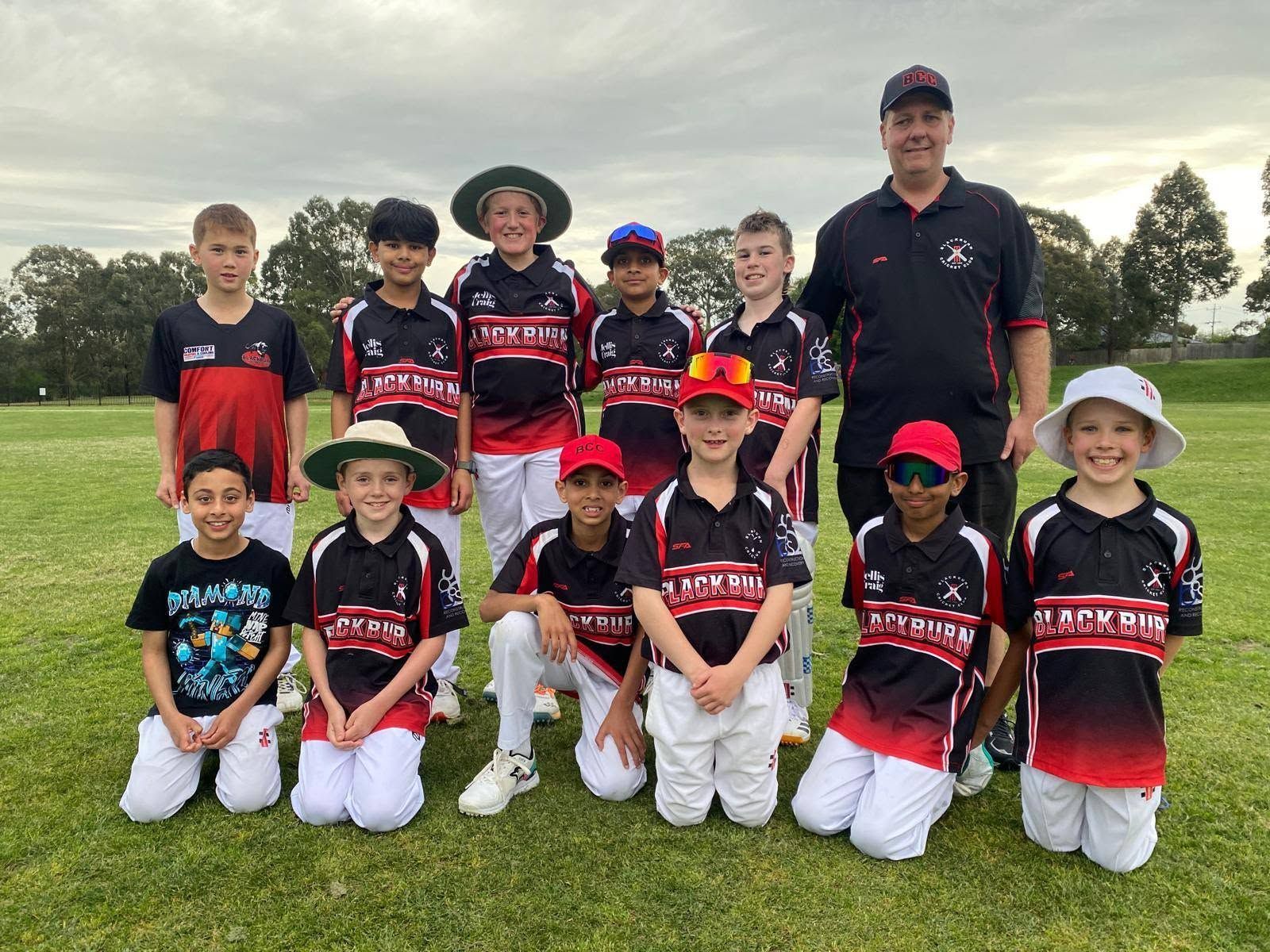 Youth cricket team and coach posing on a grassy field, wearing black and red uniforms with gloves and hats.