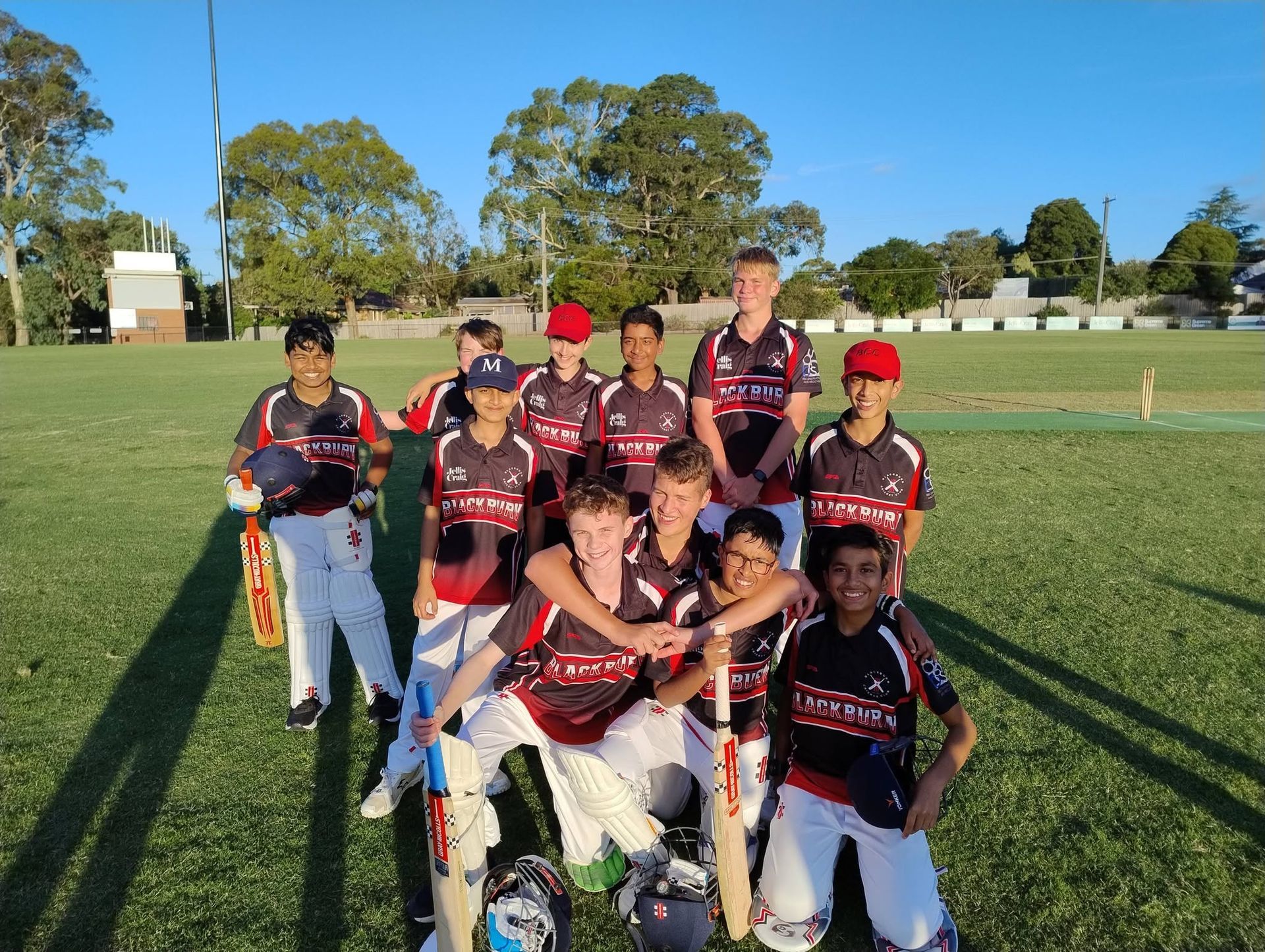 Cricket team posing on a grassy field in black and red uniforms under a clear sky