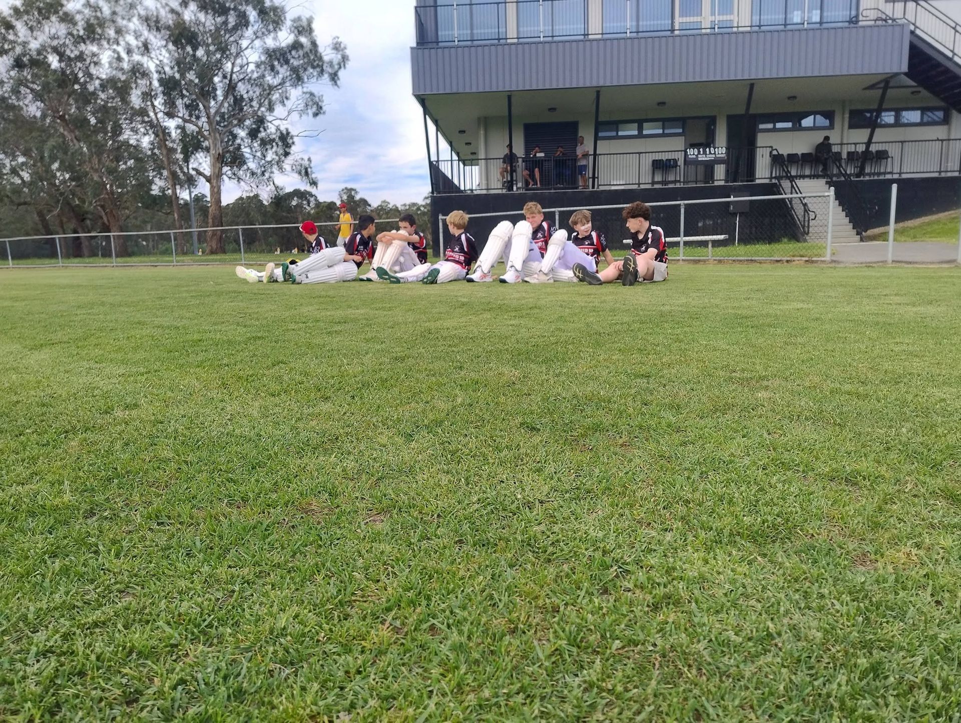 Juniors padded up, sitting on a grassy field near a stadium building, outdoors on a cloudy day