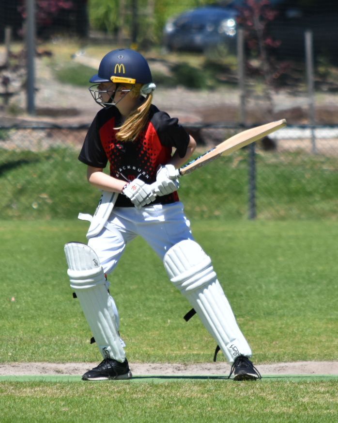 Youth female cricket player in black and white gear preparing to swing a bat on a grassy field