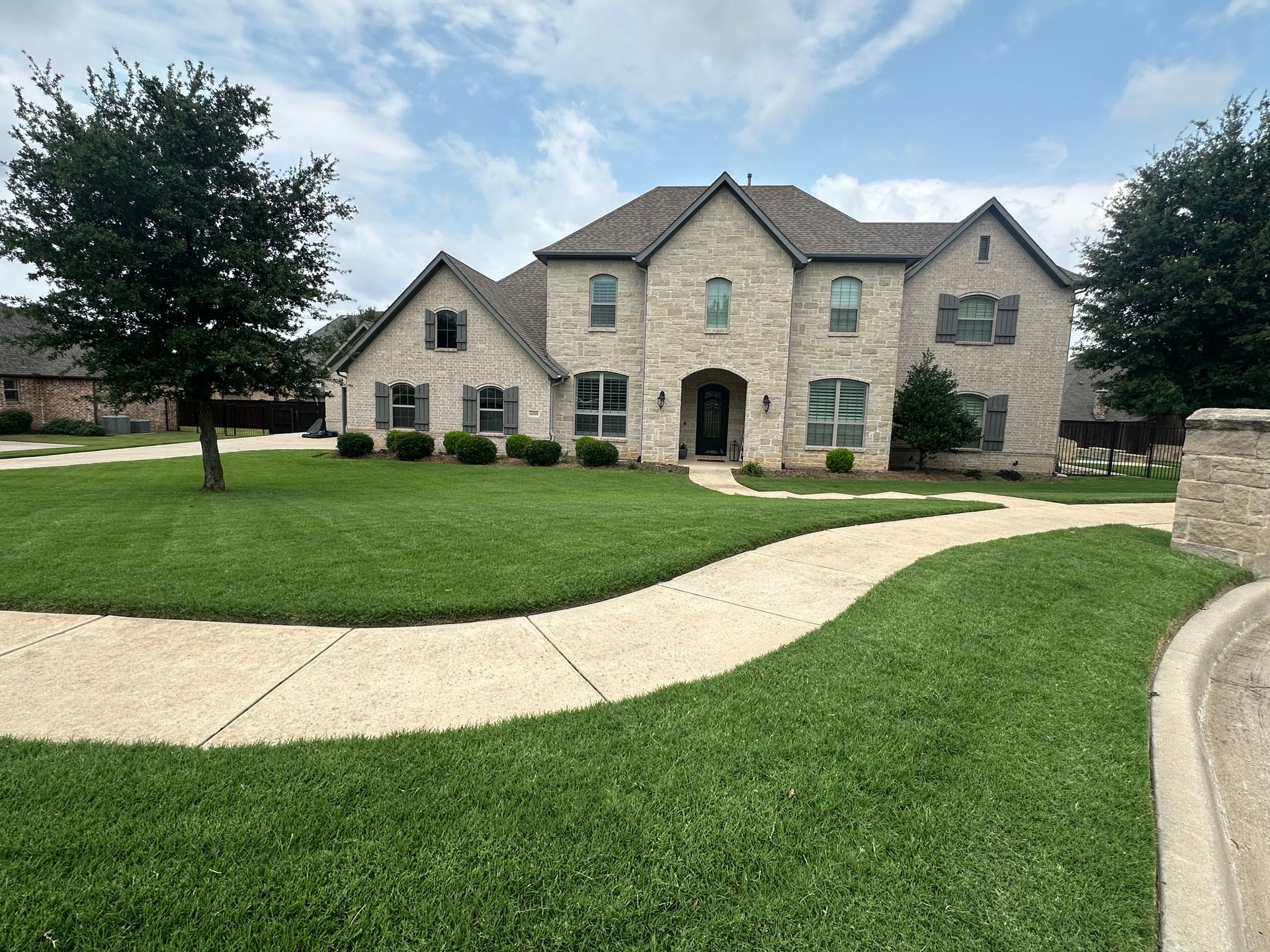 Large beige stone house with a manicured lawn, curved sidewalk, and trees in front