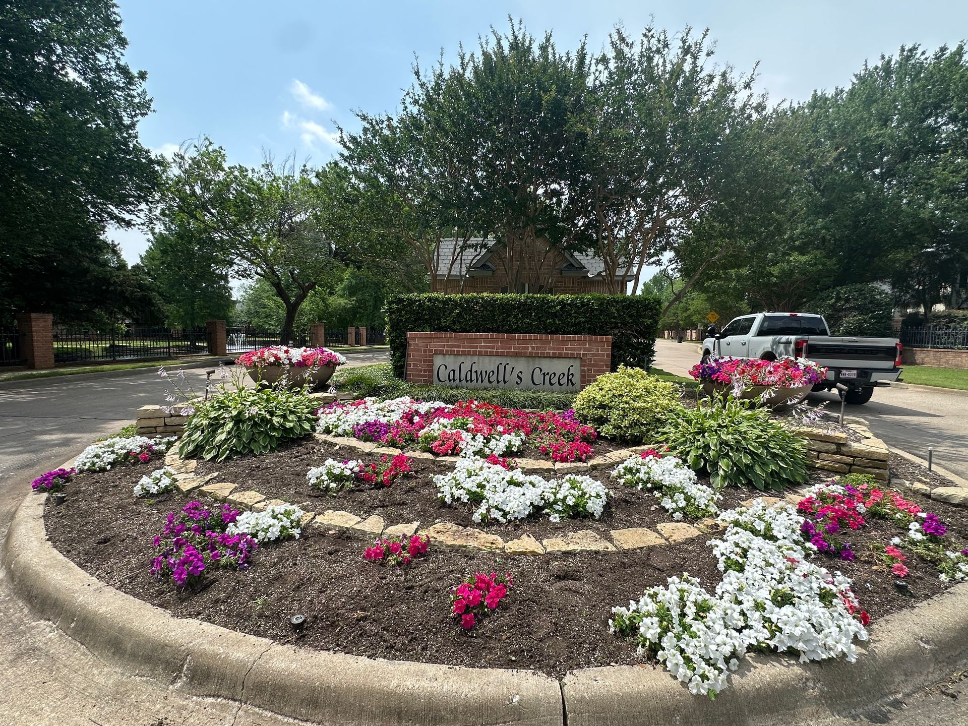 Flower bed with colorful blooms and a brick sign at a park entrance, with trees and parked vehicles in the background