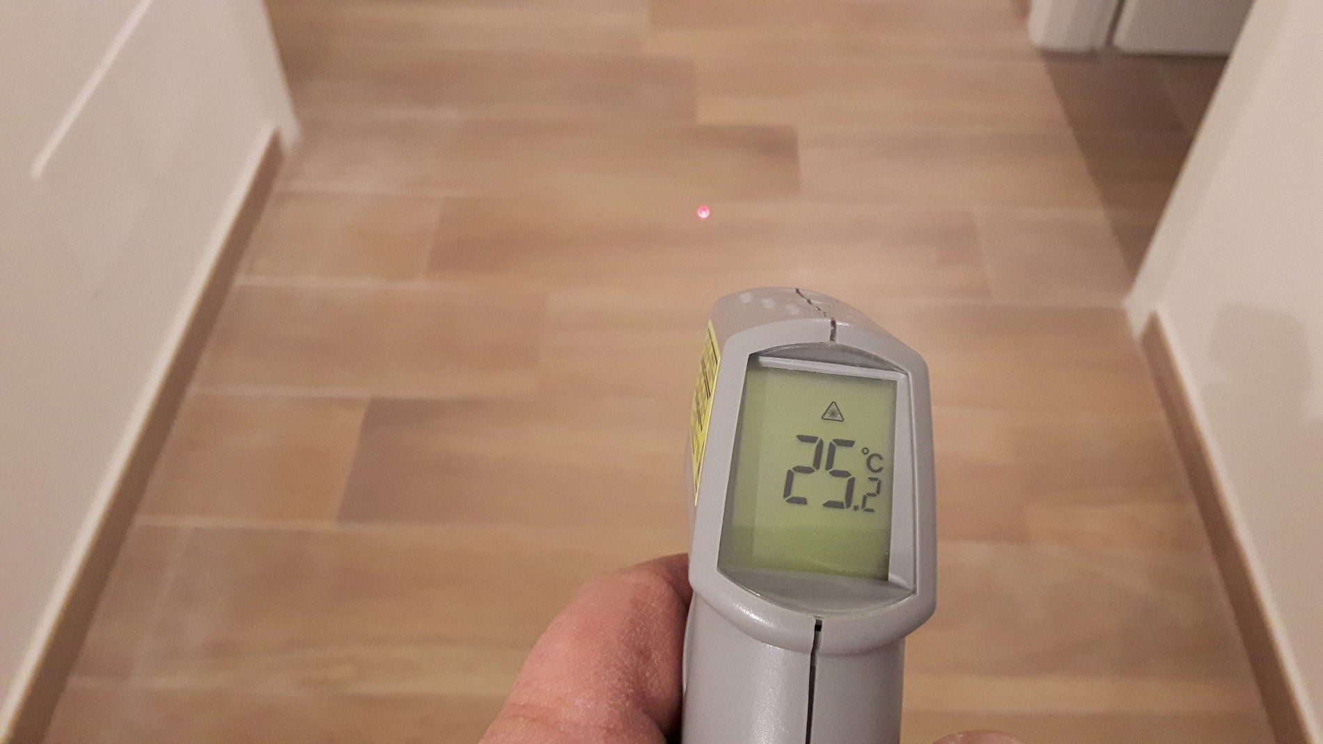 Una mano regge un termometro a infrarossi che segna 25,2 °C mentre punta un laser su un pavimento di legno chiaro in un corridoio.