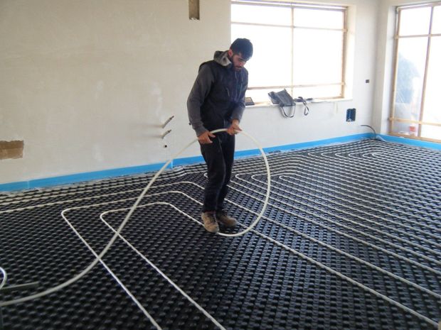 In una stanza in costruzione, una persona installa dei tubi bianchi per un sistema di riscaldamento a pavimento radiante su pannelli a griglia neri.