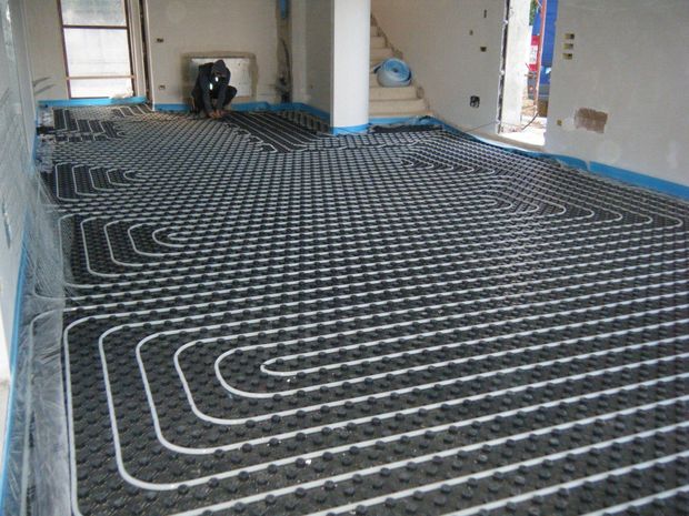 Tubi per il riscaldamento a pavimento installati su un pannello a griglia nero in una stanza in costruzione con pareti bianche.