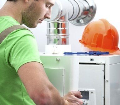 Un tecnico con una maglietta verde ispeziona un'unità di riscaldamento bianca, mentre nelle vicinanze si trova un elmetto arancione.