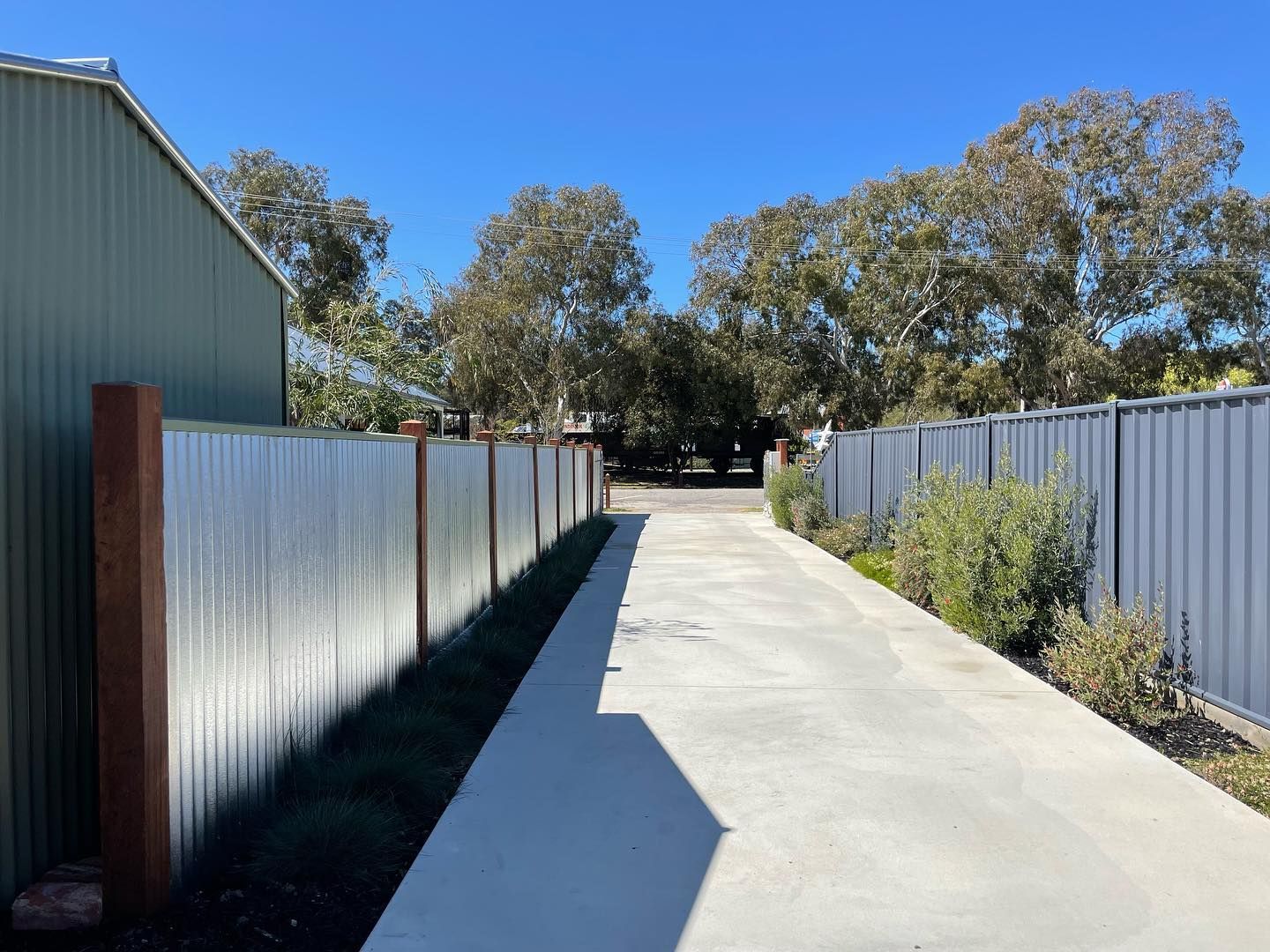 Concrete driveway between grey corrugated fences, with green building on left and trees visible at the end.