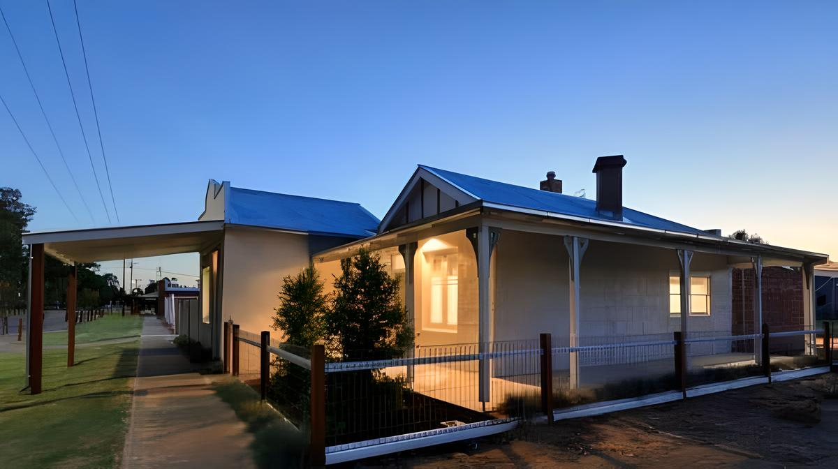 A single-story house at dusk with a porch and a fenced-in yard. The lights are on inside.
