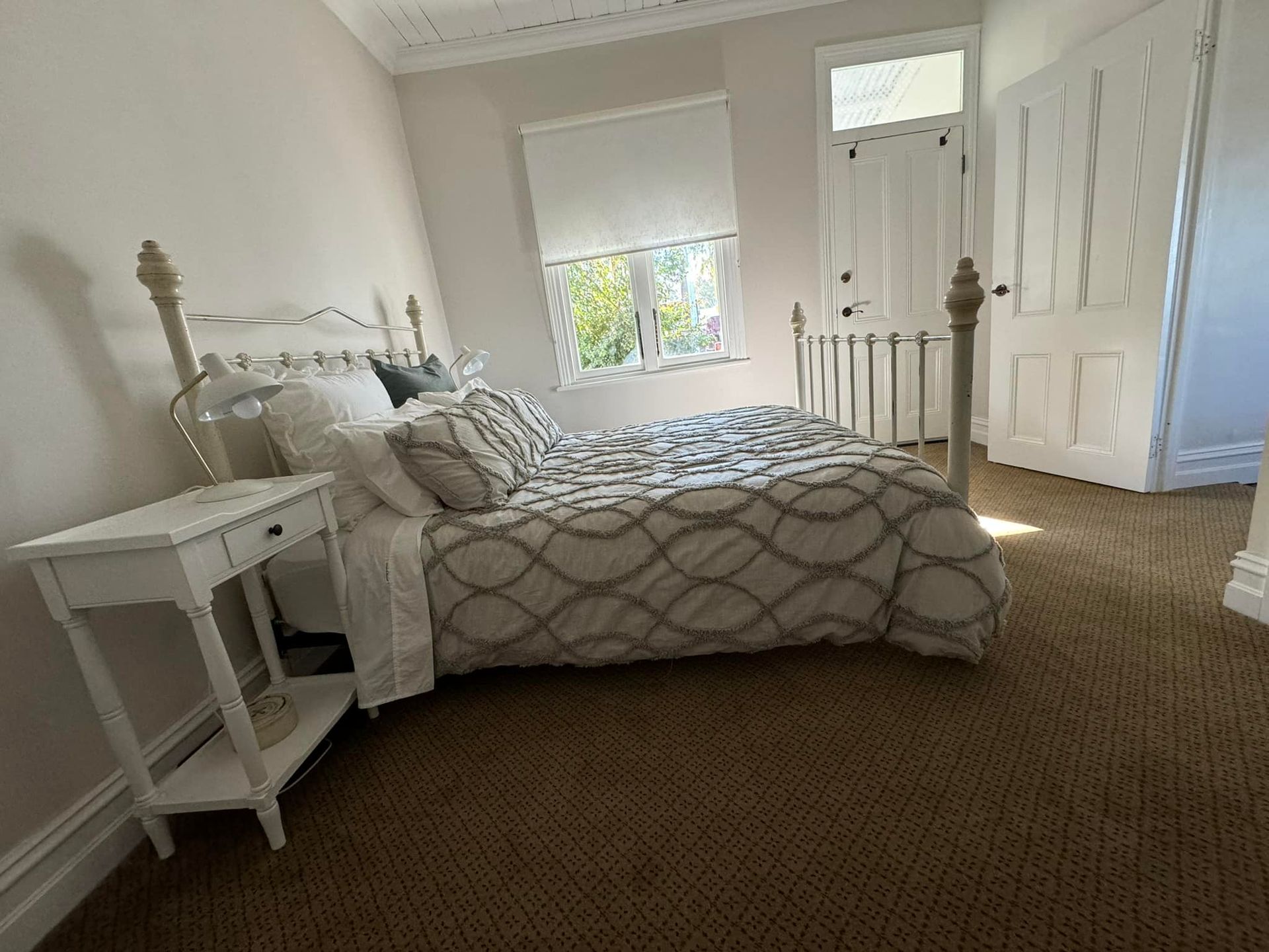 Bedroom with a bed, nightstand, and window. Soft colors, carpet flooring, and a white door.
