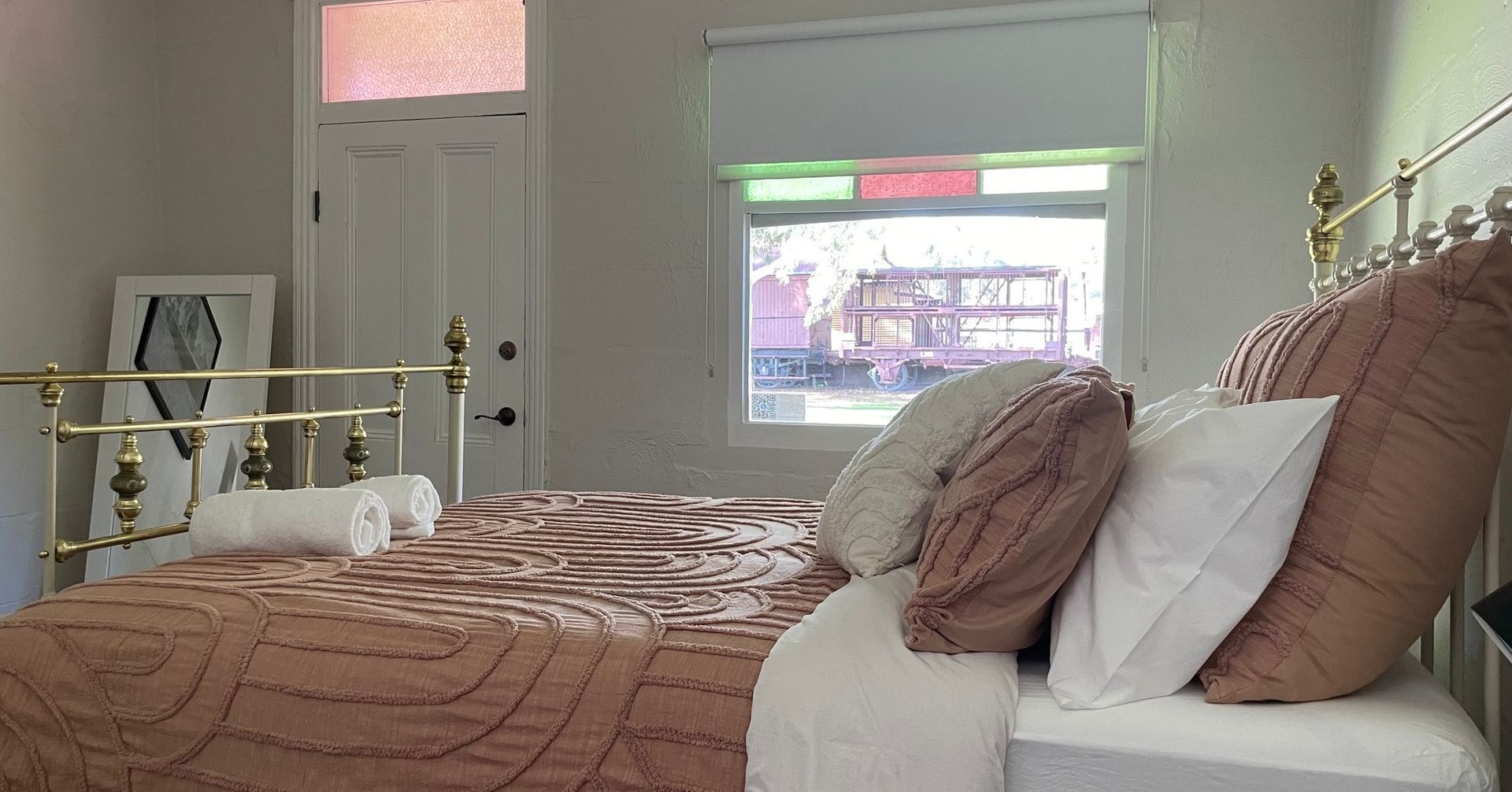Bedroom with a brass bed, window with stained glass, and a bedspread with several pillows.