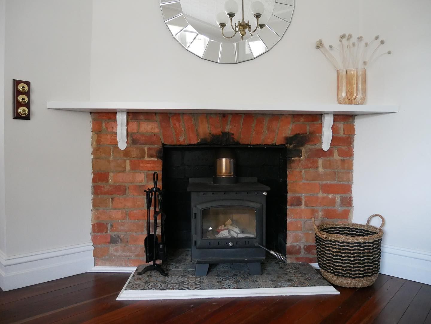 Fireplace with a wood-burning stove, brick surround, white mantel, mirror, and woven basket in a living room.