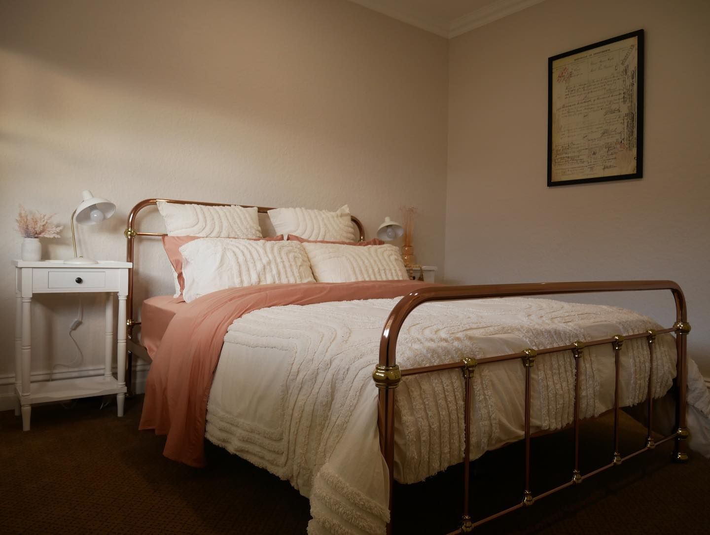 Bedroom with brass bed frame, white bedding, coral blanket, white nightstand, and abstract art.