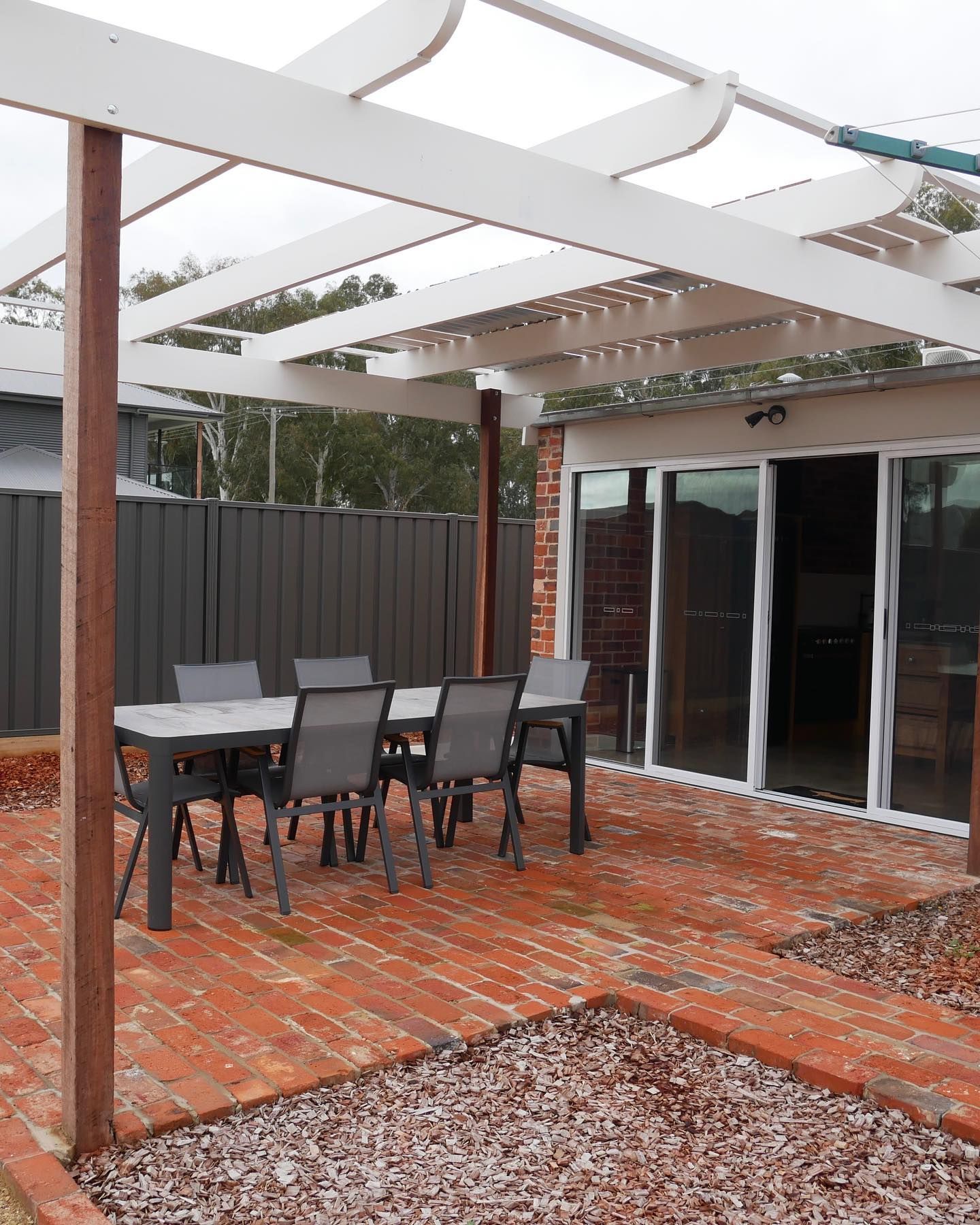 Outdoor dining area with a table and chairs under a white pergola, red brick patio.