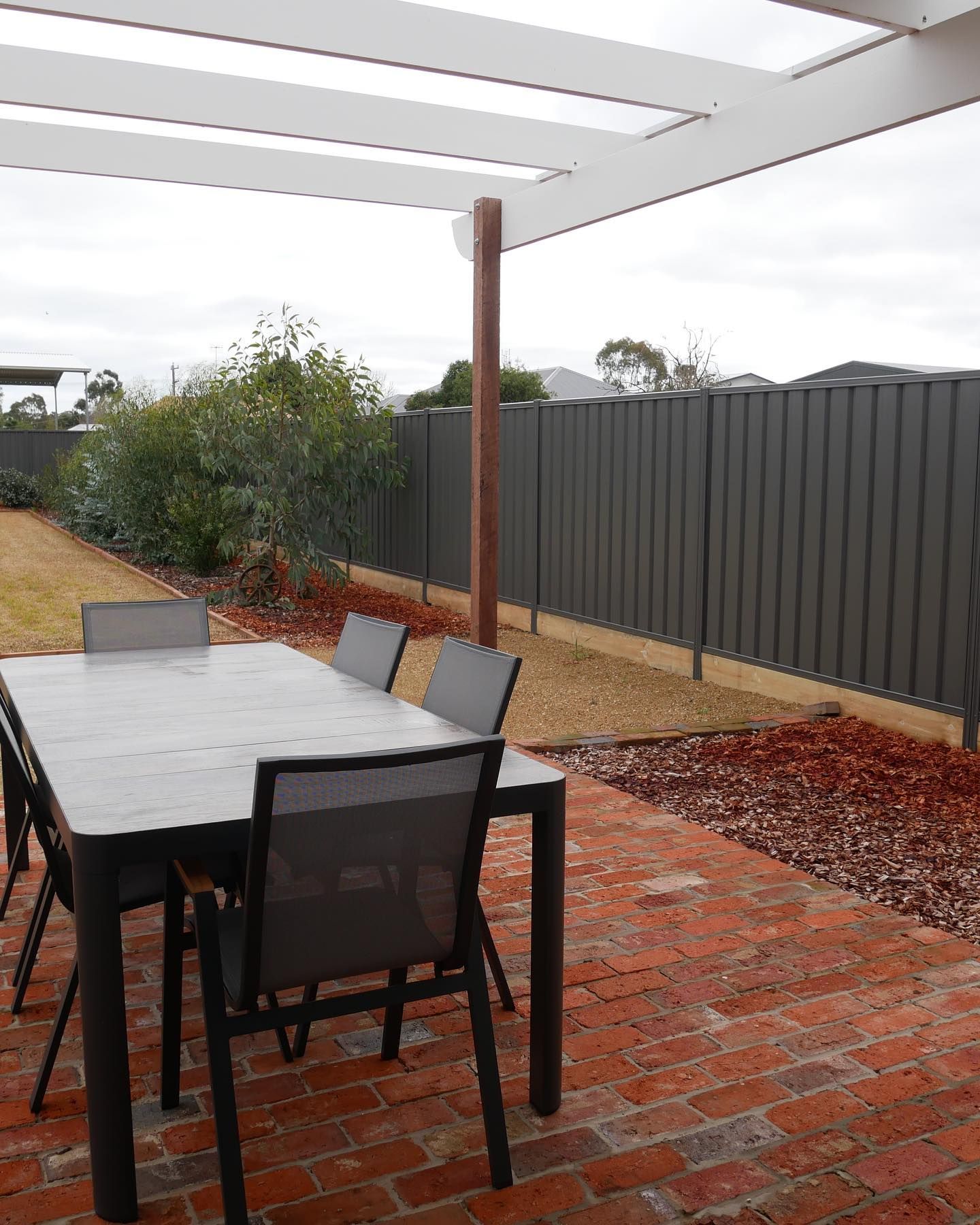 Patio with table and chairs, brick flooring, dark fence, and cloudy sky.