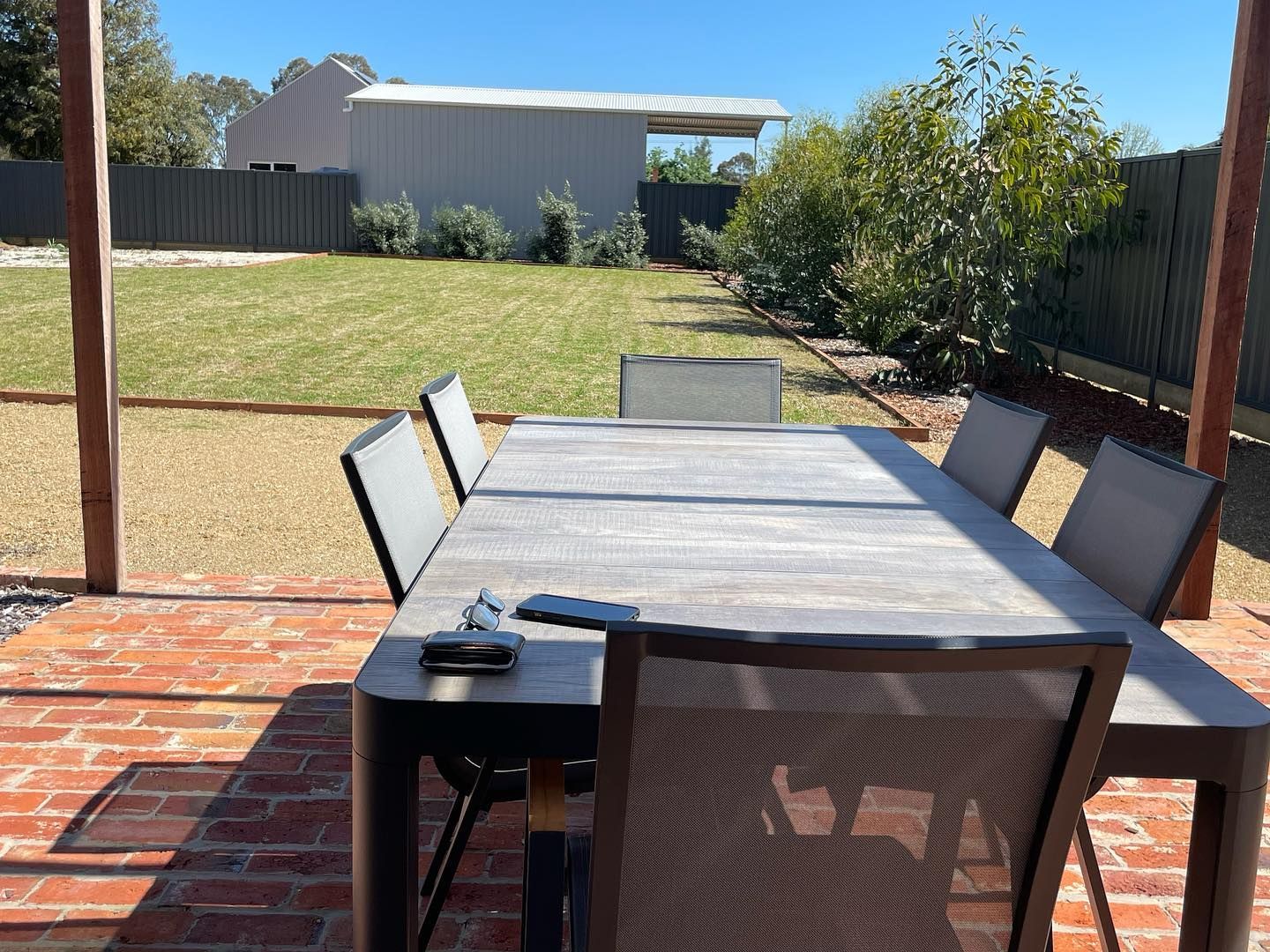 Outdoor dining set on a brick patio with a grassy yard and a shed in the background on a sunny day.