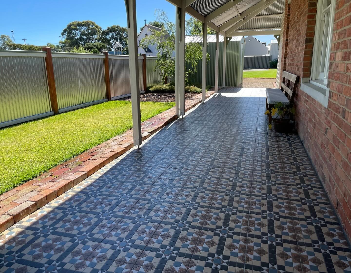 A covered porch with patterned tiles and a brick wall, next to a lawn and corrugated metal fence.