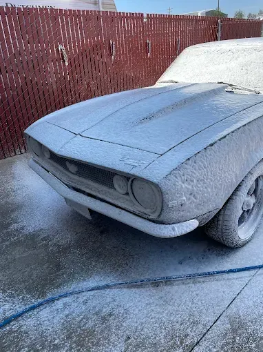 A foam-covered vintage car parked beside a red fence on a car wash service