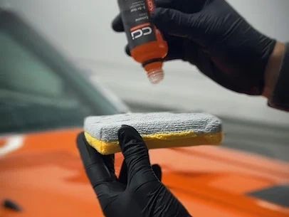 Gloved hand applying orange polish to a sponge over a car hood