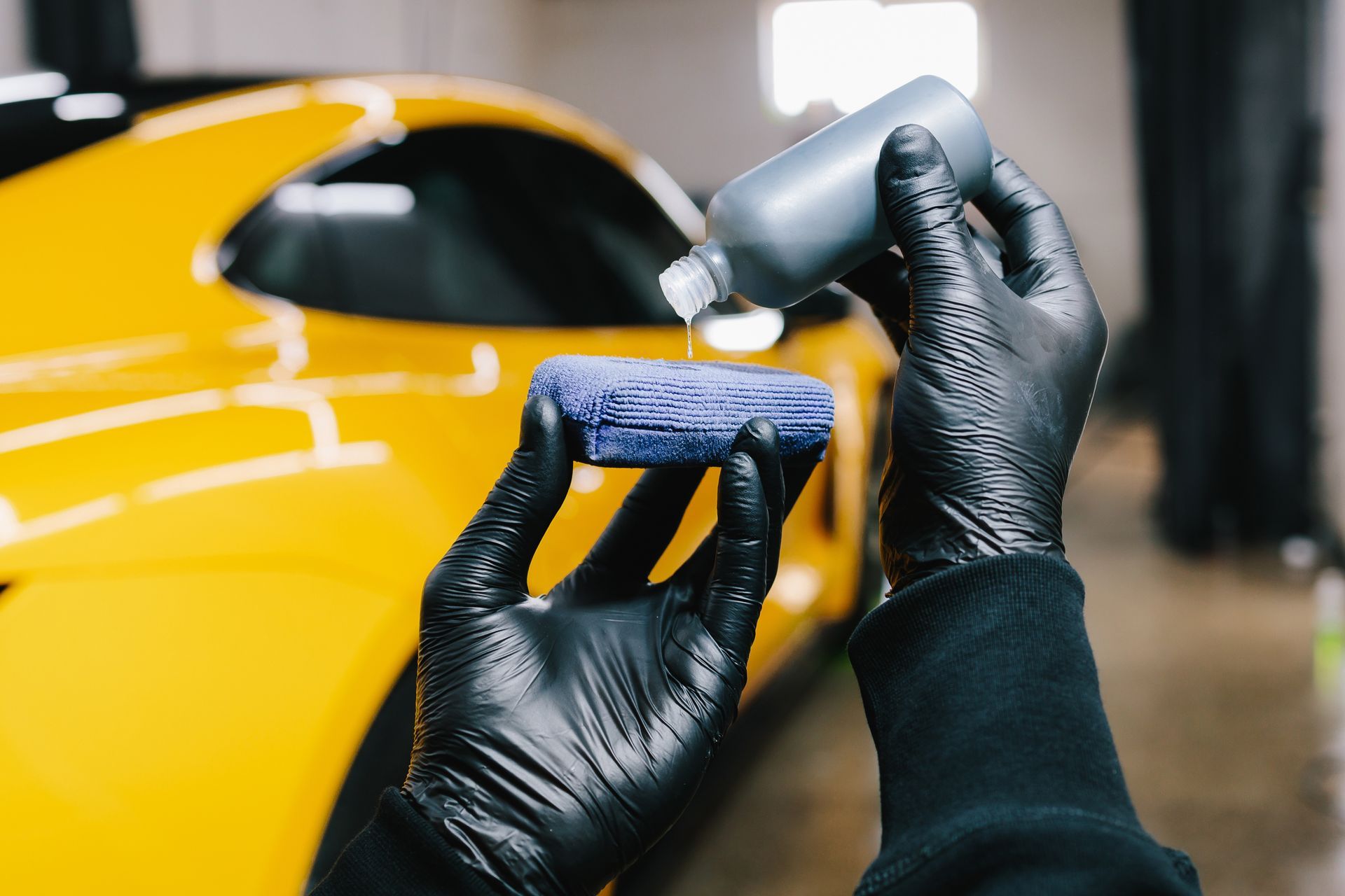 Gloved hands polishing a yellow car with a cloth and spray bottle in a garage