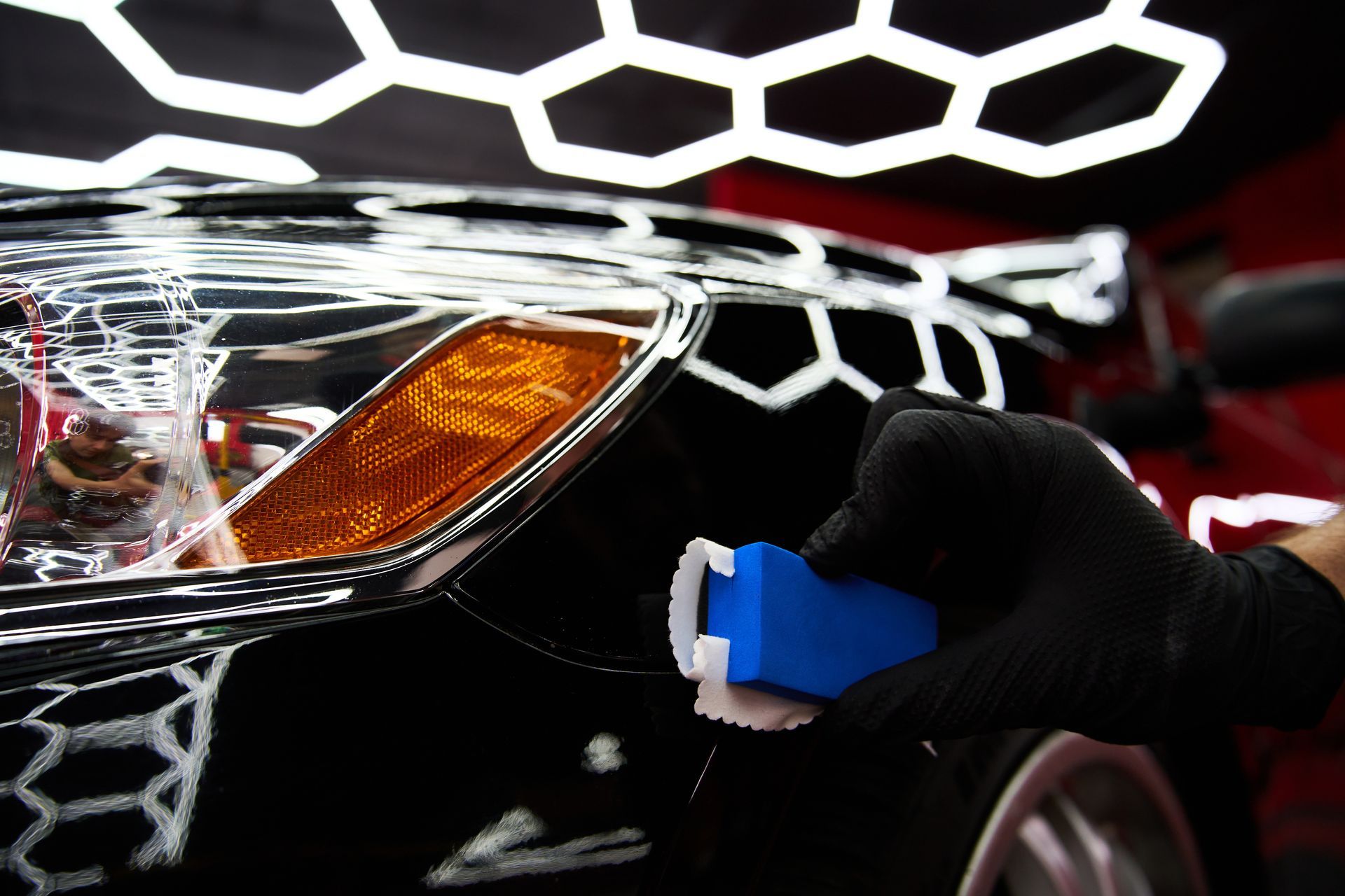 Gloved hand polishing a car’s front headlight with a blue cloth, close-up.