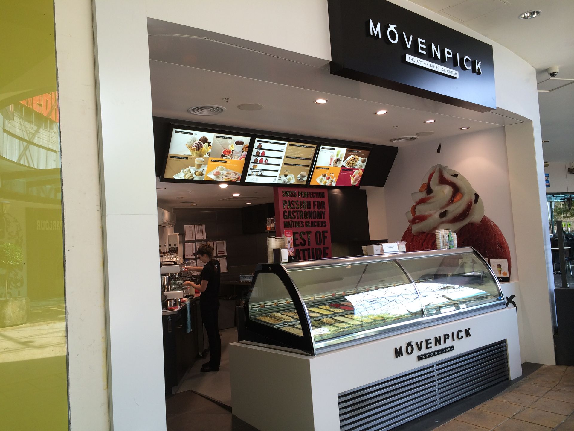 Mövenpick ice cream kiosk with illuminated menu boards and a glass display case in a mall.